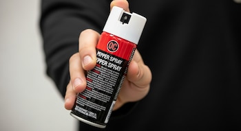 Primer plano de una mano sosteniendo un bote de gas pimienta de color rojo y blanco con la etiqueta 'PEPPER SPRAY' en un fondo oscuro.