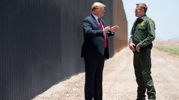 Donald Trump, vestido de traje oscuro y corbata roja, conversa con un agente de la Patrulla Fronteriza en uniforme verde junto a un muro oscuro