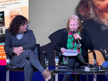 Dos mujeres sentadas en sillas en un escenario. Una con cabello oscuro y gafas, la otra con cabello claro, sostiene un micrófono y papeles. Una pantalla grande de fondo