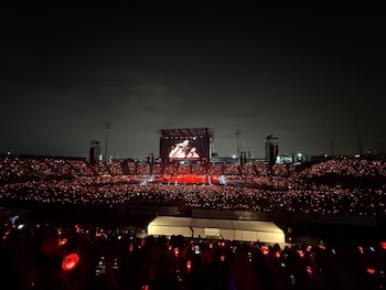 Vista nocturna de un estadio multitudinario iluminado por luces rojas de los asistentes ante un escenario con pantallas