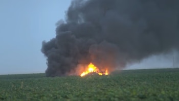 Fuego intenso con llamas anaranjadas y amarillas generando una gran columna de humo negro sobre un campo verde bajo un cielo gris claro