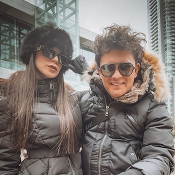 Hombre y mujer posan sonrientes con chaquetas de invierno oscuras y gafas de sol. La mujer lleva un gorro de piel, el hombre una capucha. Fondo: rascacielos