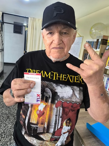 Hombre mayor con gorra y camiseta negra de Dream Theater, sostiene una entrada del concierto y hace el signo de los cuernos con su mano derecha