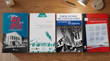 Cuatro portadas de libros colocados en fila sobre una mesa de madera junto a una lámpara y dos bolígrafos.