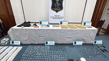 Mesa y suelo con paquetes de droga, armas de fuego, municiones, chaleco antibalas, billetes en fajos, teléfonos móviles y carteles identificadores