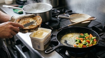 Una mano con guante unta margarina en pan tostado con un cuchillo. Una tina de margarina abierta y verduras salteándose con margarina en una sartén. Pasta hierve al fondo.