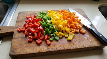 Morrones rojos, verdes, amarillos y naranjas picados sobre una tabla de madera, con un cuchillo a un lado y un fregadero al fondo.