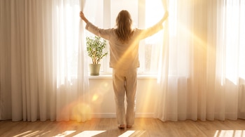 Persona de espaldas con pijama beige que abre cortinas blancas frente a una ventana iluminada con una planta en la repisa.