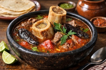 Un caldo en un cuenco de piedra con carne, dos huesos con médula, chiles chipotle secos, jitomate asado y epazote. Al lado, limones, tortillas y salsas.
