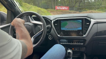 Vista interior de un vehículo con un conductor al volante; la pantalla central muestra la vista trasera y una pancarta de Isuzu se ve a través del parabrisas