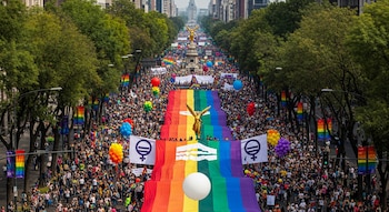 Vista aérea de una multitud en una avenida con árboles y edificios. Se ven banderas arcoíris gigantes, globos de colores, el Ángel de la Independencia y pancartas.