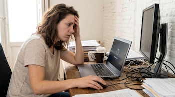 Mujer con camiseta clara y cabello castaño, apoyada en su mano, mira con preocupación la pantalla de su laptop que muestra 'Please wait...' en un escritorio de madera.