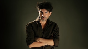 Hombre con cabello oscuro y desordenado, barba corta y camisa marrón desabrochada posa con los brazos cruzados frente a un fondo oscuro