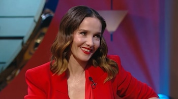 Natalia Oreiro sonriendo, con cabello ondulado castaño, blazer rojo, y labios rojos, un micrófono de solapa visible en su atuendo.