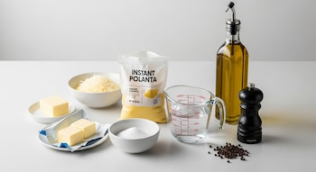 Una vista aérea de polenta instantánea, queso rallado, manteca, sal fina, agua, aceite de oliva y pimienta negra sobre una superficie blanca, listos para cocinar.
