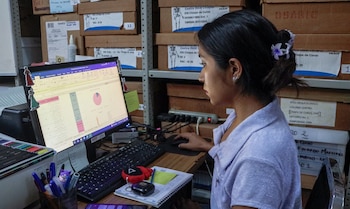 Una mujer sentada frente a un monitor con tablas de datos y gráficos, rodeada de cajas de cartón sobre estantes en un almacén