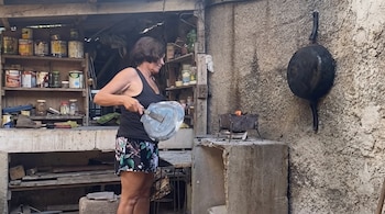 Una mujer sostiene una tapa metálica sobre un fogón encendido junto a una pared de concreto con utensilios y tarros.