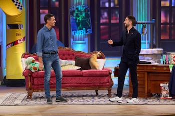 Dos hombres de pie conversan en un plató frente a un sofá rojo y un escritorio de madera sobre una alfombra