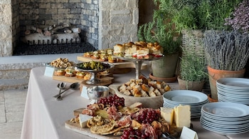 Mesa de bufé con quesos, fiambres, galletas, frutos secos, uvas, canapés, sándwiches. Platos apilados, plantas y chimenea de piedra.