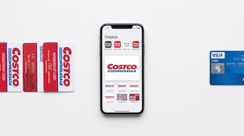Vista superior de un teléfono inteligente con pantalla de Costco entre tarjetas de membresía a la izquierda y tarjeta Visa a la derecha.
