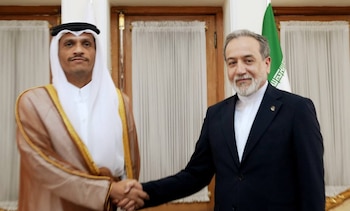 Dos hombres con vestimenta formal se estrechan la mano en una sala interior junto a la bandera de Irán y ventanas con cortinas