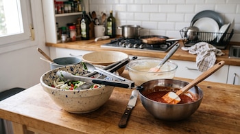 Cuencos con ingredientes, espátulas, pinzas y un cuchillo sobre una mesa de madera frente al mostrador de la cocina.