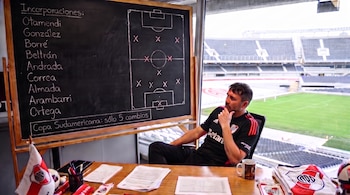 Hombre en camiseta de River Plate sentado frente a pizarra con nombres y esquema de cancha. Escritorio con bandera del club. Fondo con estadio de fútbol.