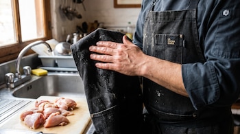 Un hombre con delantal negro se seca la mano junto a trozos de pollo crudo colocados sobre una tabla de picar cerca del fregadero.
