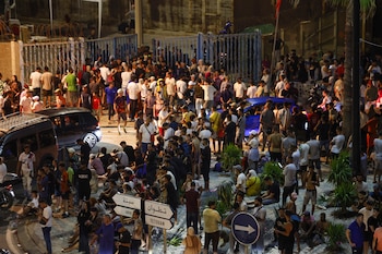 Gran multitud de personas se reúne de noche en una calle con coches, vallas con alambre de púas, palmeras y señales de tráfico a Ceuta y Tetuán