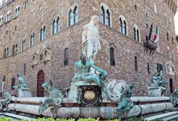Vista de la Fuente de Neptuno en Florencia, con la estatua de mármol blanco de Neptuno rodeada de figuras de bronce. Al fondo, un edificio de piedra con arcos