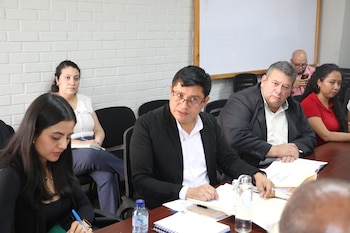 Grupo de personas sentadas alrededor de una mesa de conferencia en una sala. Varios individuos miran hacia adelante, algunos con papeles y botellas de agua en la mesa