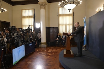 Vista general de un hombre en traje hablando desde un podio en una sala, con cámaras, periodistas y dos grandes ventanas con cortinas al fondo