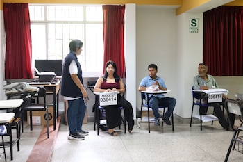 Cuatro personas en un aula. Un hombre de pie con chaleco azul y tres sentados en pupitres con carteles "MESA Nº 036414". Ventana con cortinas rojas y señal de zona sísmica