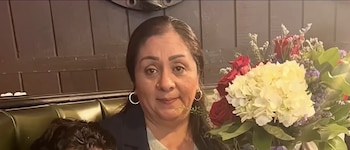 Una mujer de cabello oscuro y pendientes de aro posa junto a un ramo de rosas rojas y hortensias blancas ante una pared de madera