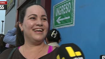 Primer plano de Annette, una mujer de piel clara y cabello oscuro, sonriendo ampliamente mientras un micrófono se acerca a ella, con un cartel verde de 'INGRESO' en el fondo azul