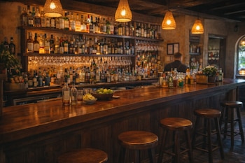 Interior de bar con barra de madera, tres taburetes, estanterías llenas de botellas y copas, y un camarero de espaldas. Lámparas colgantes.