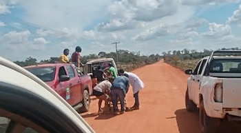 Carretera de tierra, un grupo de personas y tres vehículos (una camioneta roja, un furgón blanco y una camioneta blanca), un poste de luz, cielo nublado