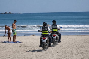 Dos agentes de policía uniformados en motocicletas blancas sobre una playa de arena. De espaldas, miran hacia el mar azul con olas. A la izquierda, un hombre y una mujer de pie en la arena.