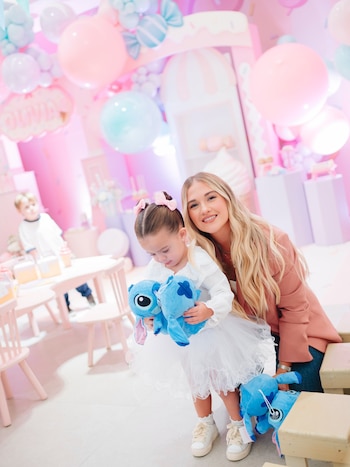 Una mujer sonríe junto a una niña con tutú blanco que sostiene peluches azules en una fiesta con globos de colores pasteles