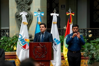 Un hombre de traje habla en un atril de madera frente a banderas institucionales, junto a un intérprete de lengua de señas