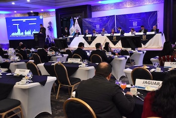 Salón con mesas redondas, manteles oscuros, rótulos de partidos políticos, autoridades en un estrado y la bandera de Guatemala