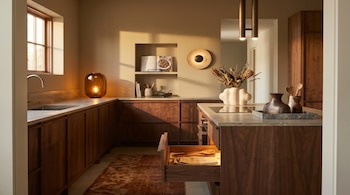 Cocina con gabinetes de madera oscura, encimeras grises claras, un fregadero de acero, y una isla central. La luz solar entra por una ventana de madera. Hay una lámpara de mesa ámbar y otra de pared circular.