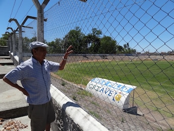 Hombre con gorra y camisa a cuadros de pie junto a un alambrado frente a una cancha de fútbol con césped verde