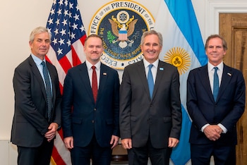 Cuatro hombres con traje y corbata posan frente a la bandera de Estados Unidos, el sello institucional y la bandera de Argentina