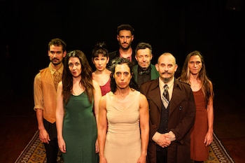 Siete personas posan en un escenario con fondo oscuro y una alfombra. Una mujer de cabello oscuro y vestido beige está al frente, con seis más detrás, en varias vestimentas