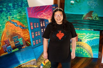Una mujer con gafas y camiseta negra posa de pie rodeada de varios cuadros al óleo con paisajes coloridos de ciudades y mar