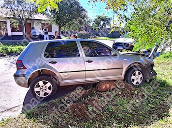 Un automóvil gris con daños en el frente sobre un montículo junto a un poste, con un patrullero y personas de pie al fondo