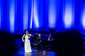 Akiko Suwanai, de pie y vestida de blanco, toca el violín en el centro del escenario, con un pianista a su derecha frente a un piano de cola bajo luces azules
