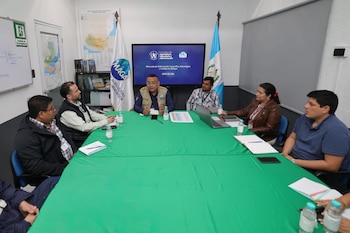 Siete personas sentadas alrededor de una mesa verde con botellas de agua y cuadernos. Detrás, una pantalla, banderas de Guatemala y del MAGA, y mapas en la pared