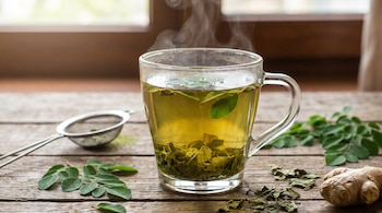 Primer plano de una taza de cristal con infusión de moringa humeante sobre una mesa de madera, junto a hojas frescas, secas, un infusor y jengibre.